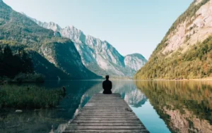 Person meditating on a jetty reaching into a calm mountain lake