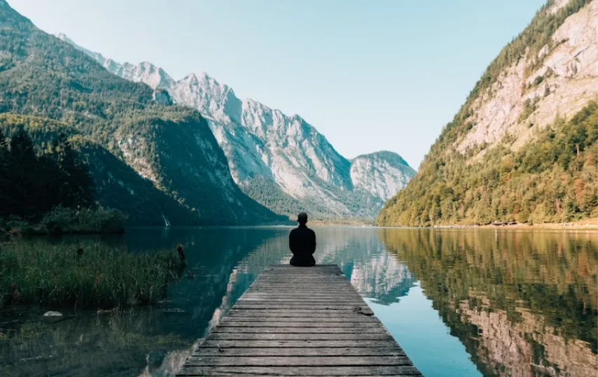Person meditating on a jetty reaching into a calm mountain lake